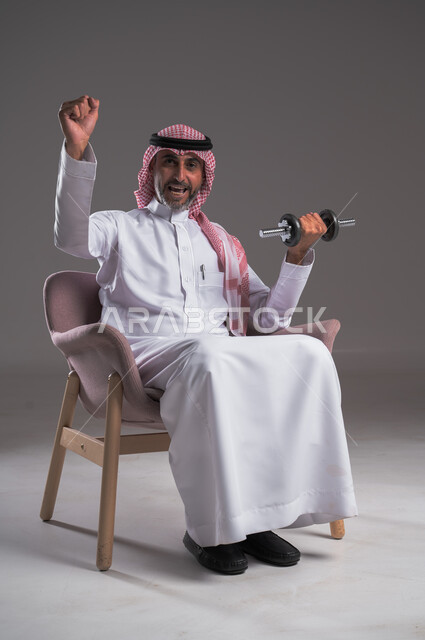 تقوية عضلات الجسم وبناؤها، تمارين رفع الأثقال والأوزان الحديدية الثقيلة، القوة والتحدي والتحفيز لأسلوب حياة صحي، بورتريه لرجل عربي خليجي سعودي مبتسم يرتدي الثوب التقليدي والشماغ يجلس على كرسي مريح ويمارس التمارين الرياضية بواسطة الدامبلز، خلفية رمادية