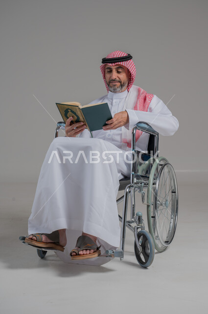 مفهوم الاجتهاد في العبادة، قراءة كتاب الله وحفظه وتجويده، تلاوة المصحف الشريف، بورتريه لرجل عربي خليجي سعودي مقعد يرتدي الثوب التقليدي والشماغ يجلس على كرسي متحرك يحمل بيده القرآن، صورة لكامل الجسم، خلفية رمادية