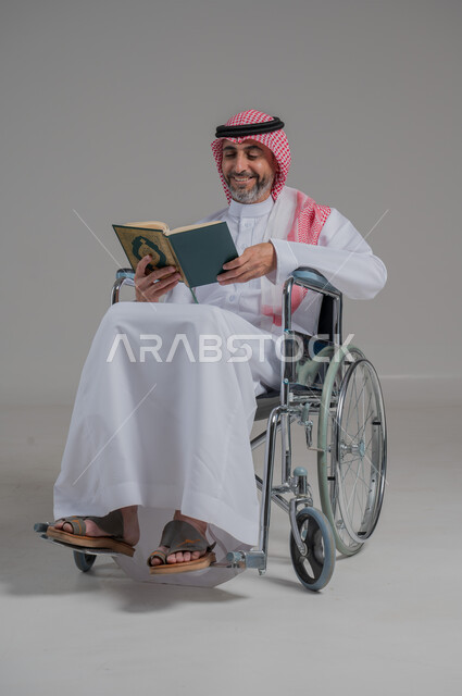 تلاوة المصحف الشريف، مفهوم الاجتهاد في العبادة، قراءة كتاب الله وحفظه وتجويده، بورتريه لرجل عربي خليجي سعودي مقعد يرتدي الثوب التقليدي والشماغ يجلس على كرسي متحرك يحمل بيده القرآن، صورة لكامل الجسم، خلفية رمادية
