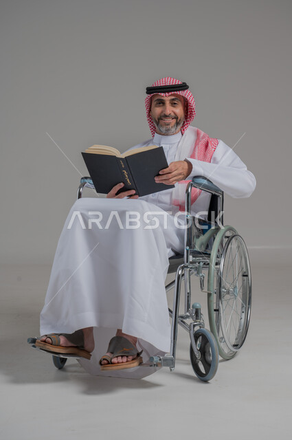 أسلوب حياة ذوي الهمم، دعم المجتمع السعودي لهوايات أصحاب الاحتياجات الخاصه، الاستمتاع بممارسة هواية المطالعة، بورتريه لرجل عربي خليجي سعودي مقعد يرتدي الثوب التقليدي والشماغ يجلس على كرسي متحرك يقوم بقراءة كتاب، صورة لكامل الجسم، خلفية رمادية
