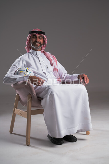 Enthusiasm and interaction with the team, different sports clubs, gestures of passion and encouragement for the team, watching and following a football match, portrait of a smiling Saudi Arabian Gulf man wearing traditional thobe and shemagh sitting on a comfortable chair holding the ball, looking at the camera with happy expressions, gray background