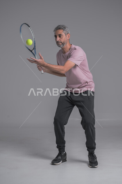 Recreational activities and youth sports, training to master the game of tennis, performing sports exercises, portrait of an elderly Saudi Arabian Gulf man wearing sports clothing and playing with a ball and racket, gestures indicating enthusiasm and engagement with exercise, full body image, gray background