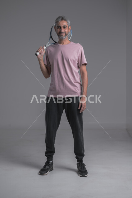 Recreational activities and youth sports, training to master the game of tennis, performing sports exercises, portrait of an elderly Saudi Arabian Gulf man wearing sports clothing and playing with a ball and racket, gestures indicating enthusiasm and engagement with exercise, full body image, gray background