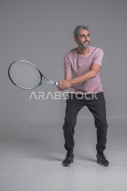 التدرب على اتقان لعبة التنس, أنشطة ترفيهية و رياضات شبابية، إجراء تمرينات رياضة، بورتريه لرجل كبير بالسن عربي خليجي سعودي يرتدي ملابس رياضية و يلعب بالكرة والمضرب، إيماءات تدل على الحماس والاندماج بالتمرين، صورة الجسم بالكامل، خلفية رمادية