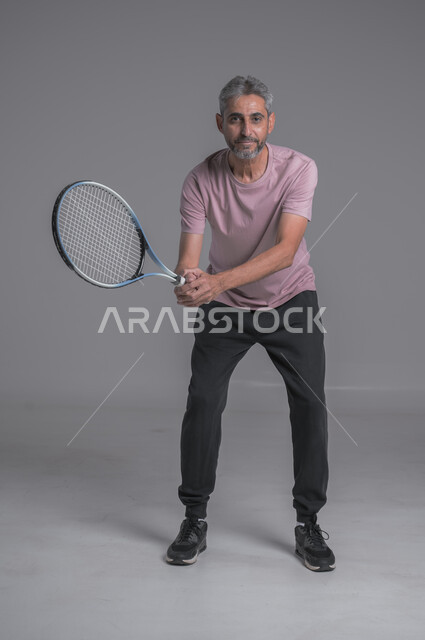 التدرب على اتقان لعبة التنس, أنشطة ترفيهية و رياضات شبابية، إجراء تمرينات رياضة، بورتريه لرجل كبير بالسن عربي خليجي سعودي يرتدي ملابس رياضية و يلعب بالكرة والمضرب، إيماءات تدل على الحماس والاندماج بالتمرين، صورة الجسم بالكامل، خلفية رمادية
