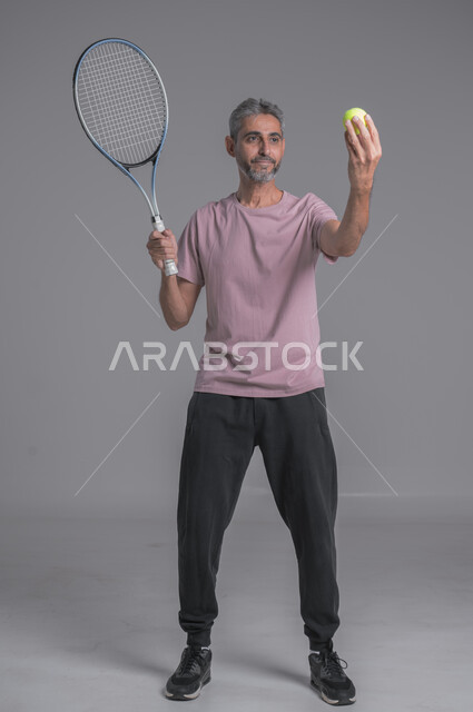 التدرب على اتقان لعبة التنس, أنشطة ترفيهية و رياضات شبابية، إجراء تمرينات رياضة، بورتريه لرجل كبير بالسن عربي خليجي سعودي يرتدي ملابس رياضية و يلعب بالكرة والمضرب، إيماءات تدل على الحماس والاندماج بالتمرين، صورة الجسم بالكامل، خلفية رمادية