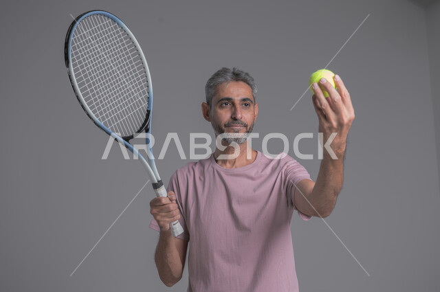 التدرب على اتقان لعبة التنس, أنشطة ترفيهية و رياضات شبابية، إجراء تمرينات رياضة، بورتريه لرجل كبير بالسن عربي خليجي سعودي يرتدي ملابس رياضية و يلعب بالكرة والمضرب، إيماءات تدل على الحماس والاندماج بالتمرين، صورة الجسم بالكامل، خلفية رمادية