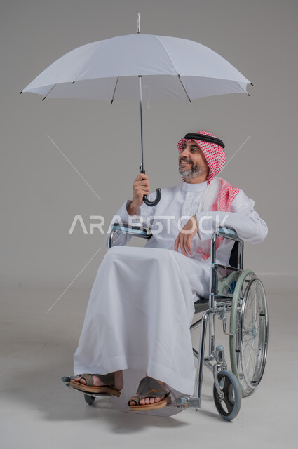 التظلل من أشعة الشمس، دمج ذوي الهمم بالمجتمع السعودي، الحماية من الأمطار في الشتاء، بورتريه لرجل عربي خليجي سعودي مقعد يرتدي الثوب التقليدي والشماغ يجلس على كرسي متحرك يحمل بيده مظلة، صورة لكامل الجسم، خلفية رمادية