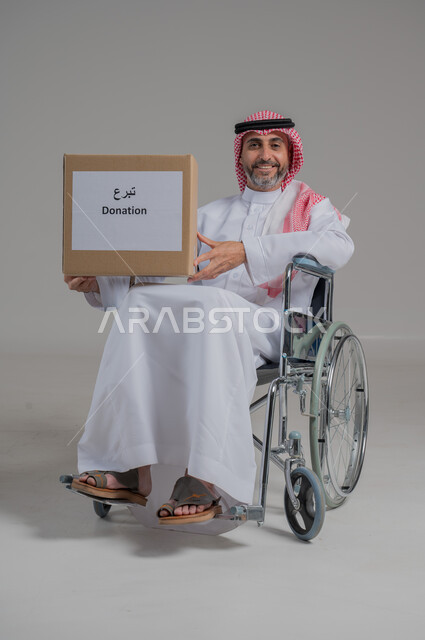 Humanitarian aid and donations, Islamic charity, feeling for others and helping them, portrait of a disabled Saudi Arabian Gulf man wearing traditional thobe and shemagh sitting in a wheelchair holding a donation box, full body image, gray background