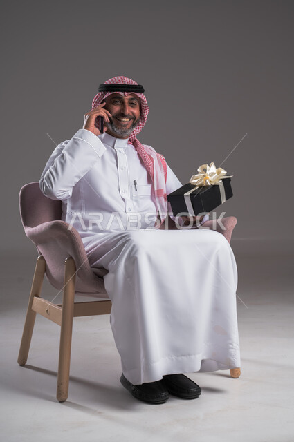 التواصل مع الأصدقاء والاقارب، سماع أخبار سارَة عبر الهاتف المحمول، بورتريه لرجل عربي خليجي سعودي مبتسم يرتدي الثوب التقليدي والشماغ يجلس على كرسي مريح يقوم بإجراء مكالمة هاتفية ويحمل بيده علبة هدية بإيماءات السرور، أعياد ومفاجآت ومناسبات سعيدة، خلفية رمادية