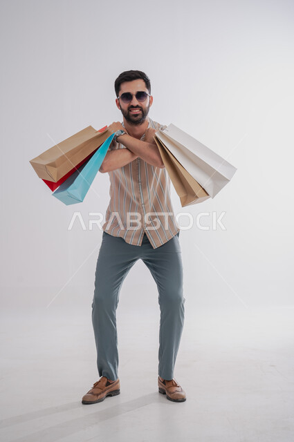 الاستفادة من عروض وتخفيضات الجمعة البيضاء، تعابير الحماس للتسوق واقتناء المستلزمات والهدايا،  بورتريه لرجل عربي خليجي سعودي مبتسم يحمل بيده أكياس ورقية ملونة ينظر ويرتدي نظارات شمسية، صورة الجسم بالطول الكامل، خلفية بيضاء