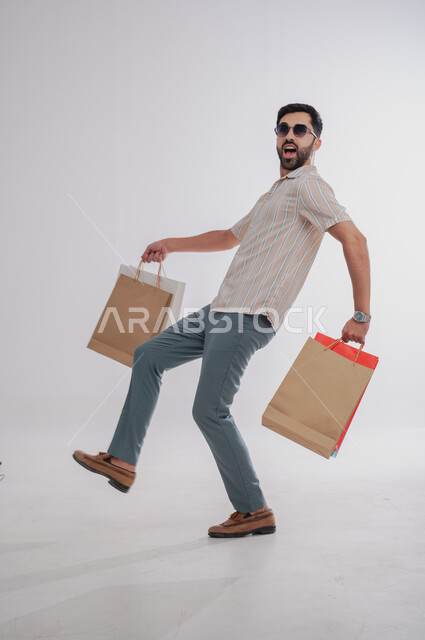 Taking advantage of Black Friday offers and sales, expressions of enthusiasm for shopping and purchasing supplies and gifts, a portrait of a smiling Saudi Arabian Gulf man holding colored paper bags in his hand, looking at and wearing sunglasses, full-length body portrait, white background.