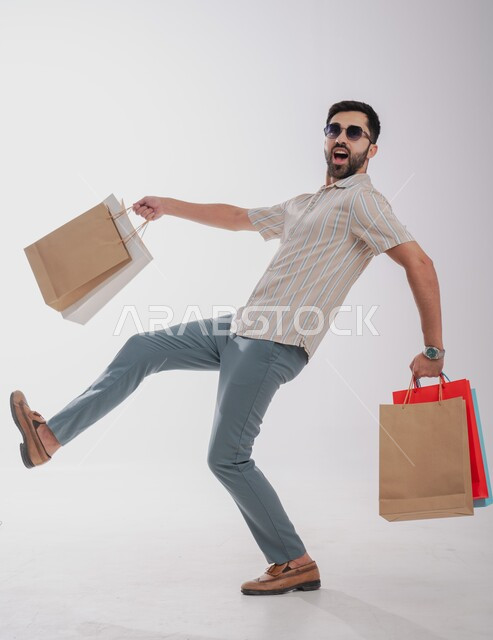 الاستفادة من عروض وتخفيضات الجمعة البيضاء، تعابير الحماس للتسوق واقتناء المستلزمات والهدايا،  بورتريه لرجل عربي خليجي سعودي مبتسم يحمل بيده أكياس ورقية ملونة ينظر ويرتدي نظارات شمسية، صورة الجسم بالطول الكامل، خلفية بيضاء