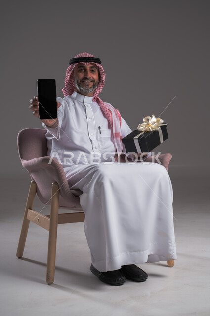 أعياد ومفاجآت ومناسبات سعيدة، بورتريه لرجل عربي خليجي سعودي مبتسم يرتدي الثوب التقليدي والشماغ يجلس على كرسي مريح يحمل بيده علبة هدية وموبايل، عرض شاشة سوداء فارغة على الهاتف المحمول، مفهوم الإهداء والتقدير، ايماءات تدل على السعادة والسرور، خلفية رمادية