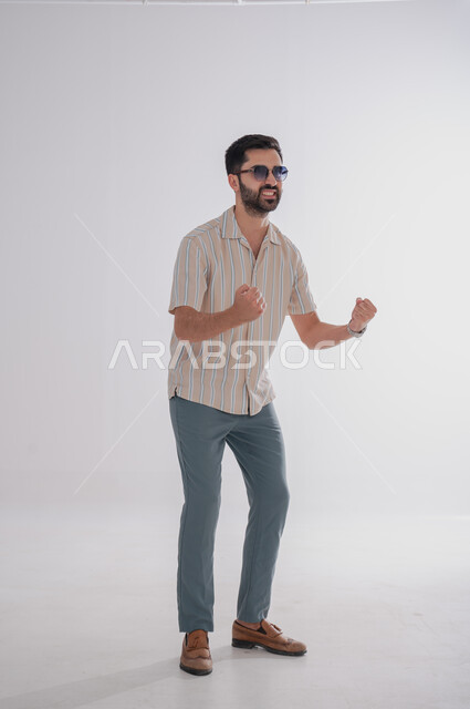 Gestures of happiness and joy, expressions of enthusiasm and pleasure, portrait of a smiling Saudi Gulf Arab man wearing sunglasses, full-length body portrait, white background