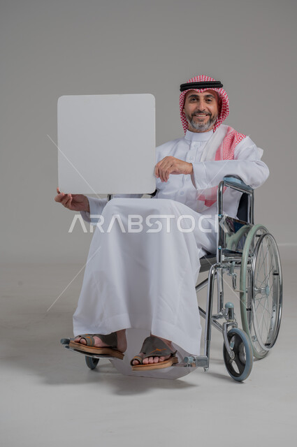 إعلانات وعروض تسويقية، موك اب مربع أبيض، استخدام لوحات توضيحية ووسائل تعليمية، بورتريه لرجل عربي خليجي سعودي مقعد يرتدي الثوب التقليدي والشماغ يجلس على كرسي متحرك يحمل بيده لوحة بيضاء فارغة، صورة لكامل الجسم، خلفية رمادية
