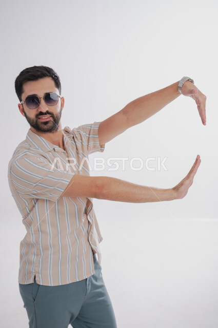 Looking at the camera and identifying a specific thing with gestures of pleasure, portrait of a smiling Saudi Gulf Arab man wearing sunglasses, white background