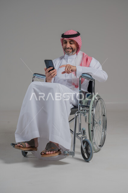 استخدام التكنولوجيا و التقنيات الحديثة، أسلوب حياة ذوي الهمم، الدردشة والتواصل مع الأهل والأصدقاء، بورتريه لرجل عربي خليجي سعودي مقعد يرتدي الثوب التقليدي والشماغ يجلس على كرسي متحرك يحمل بيده الهاتف المحمول، صورة لكامل الجسم، خلفية رمادية