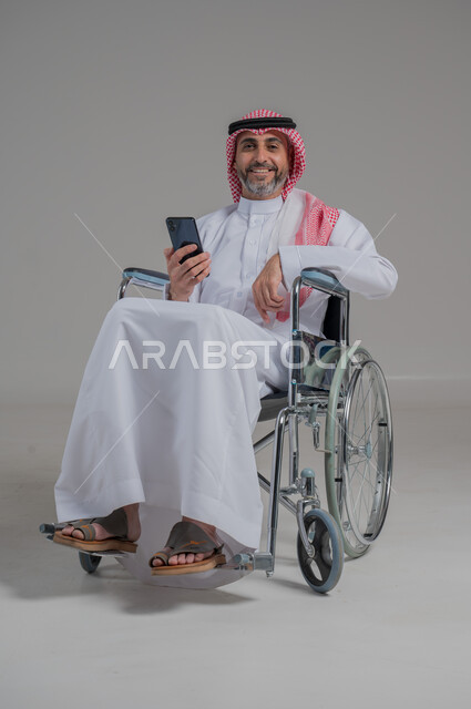 Lifestyle of people with disabilities, use of technology and modern techniques, chatting and communicating with family and friends, portrait of a disabled Saudi Arabian Gulf man wearing traditional thobe and shemagh sitting on a wheelchair holding a mobile phone in his hand, full body image, gray background