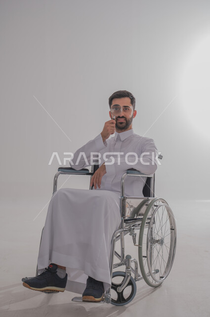 البحث والتدقيق بإيماءات الحماس والبشاشة، بورتريه لشاب عربي خليجي سعودي من ذوي الاحتياجات الخاصة يرتدي الثوب التقليدي يجلس على كرسي متحرك يمسك بيده عدسة مكبرة، مفهوم توضيح الرؤية والفحص والاستكشاف، الجسم بالطول الكامل، خلفية رمادية