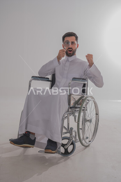 البحث والتدقيق بإيماءات الحماس والبشاشة، بورتريه لشاب عربي خليجي سعودي من ذوي الاحتياجات الخاصة يرتدي الثوب التقليدي يجلس على كرسي متحرك يمسك بيده عدسة مكبرة، مفهوم توضيح الرؤية والفحص والاستكشاف، الجسم بالطول الكامل، خلفية رمادية