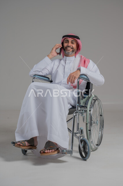 Making phone calls, gestures of pleasure and happiness, using modern technical devices, communicating with family and friends, portrait of a disabled Saudi Arabian Gulf man wearing traditional thobe and shemagh sitting on a wheelchair holding a mobile phone in his hand, full body image, gray background
