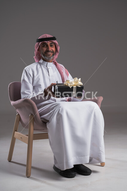 Congratulations and blessings for success and joy, portrait of a smiling Saudi Arabian Gulf man wearing traditional thobe and shemagh sitting on a comfortable chair holding a gift box in his hand, holidays, surprises and happy occasions, looking at the camera with gestures of happiness and pleasure, gray background