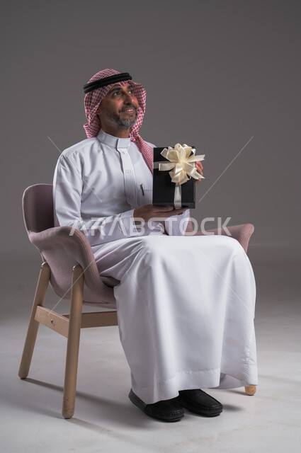 زيادة روابط المحبة بالإهداء والتقدير, التهنئات والمباركات بالنجاح والأفراح, النظر إلى شيء ما بإيماءات السعادة والسرور، بورتريه لرجل عربي خليجي سعودي مبتسم يرتدي الثوب التقليدي والشماغ يجلس على كرسي مريح يحمل بيده علبة هدية، أعياد ومناسبات سعيدة، خلفية رمادية