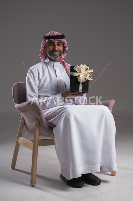 النظر الى الكاميرا بايماءات السعادة والسرور، التهنئات والمباركات بالنجاح والأفراح, بورتريه لرجل عربي خليجي سعودي مبتسم يرتدي الثوب التقليدي والشماغ يجلس على كرسي مريح يحمل بيده علبة هدية ، أعياد ومفاجآت ومناسبات سعيدة، خلفية رمادية