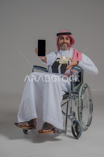 Showing a blank black screen, Saudi community support for people with disabilities, happy occasions and holidays, portrait of a disabled Saudi Arabian Gulf man wearing traditional thobe and shemagh sitting on a wheelchair holding a gift box and mobile phone, the feeling of love and appreciation for the disabled, full body image, gray background