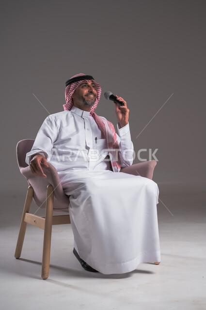 الاستمتاع بأداء المواهب الصوتية، بورتريه لرجل عربي خليجي سعودي مبتسم يرتدي الثوب التقليدي والشماغ يجلس على كرسي مريح يحمل بيده المايك، إيماءات وتعابير الاندماج والتفاعل مع الموسيقا، تسجيل مقطع صوتي غنائي، تقديم العروضات المسرحية، خلفية رمادية