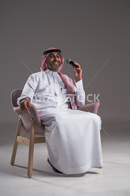 تسجيل مقطع صوتي غنائي، تقديم العروضات المسرحية، الاستمتاع بأداء المواهب الصوتية،  بورتريه لرجل عربي خليجي سعودي مبتسم يرتدي الثوب التقليدي والشماغ يجلس على كرسي مريح ينظر الى الكاميرا ويحمل بيده المايك، تعابير الاندماج والتفاعل مع الموسيقا، خلفية رمادية