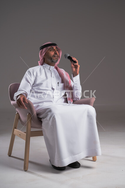 تقديم العروضات المسرحية، الاستمتاع بأداء المواهب الصوتية، بورتريه لرجل عربي خليجي سعودي مبتسم يرتدي الثوب التقليدي والشماغ يجلس على كرسي مريح يحمل بيده المايك، إيماءات وتعابير الاندماج والتفاعل مع الموسيقا، تسجيل مقطع صوتي غنائي، خلفية رمادية
