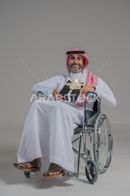 Saudi society's interest and support for people with disabilities, happy occasions and holidays, portrait of a disabled Saudi Gulf Arab man wearing traditional thobe and shemagh sitting in a wheelchair holding a gift box, the feeling of love and appreciation for the disabled, full body image, gray background