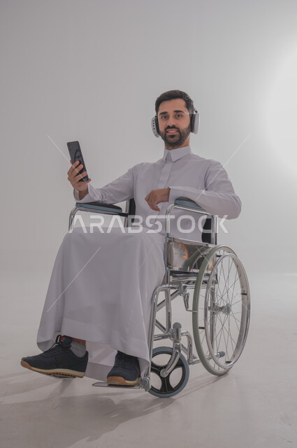 استخدام التقنيات الحديثة والتكنولوجيا المتطورة، بورتريه لشاب عربي خليجي سعودي من ذوي الاحتياجات الخاصة يرتدي الثوب التقليدي يجلس على كرسي متحرك يضع سماعات الأذن ويمسك بيده الهاتف المحمول، الاستماع إلى الموسيقا المفضلة والتمتع بقضاء أوقات الفراغ، خلفية رمادية