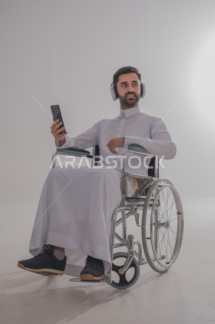 استخدام التقنيات الحديثة والتكنولوجيا المتطورة، بورتريه لشاب عربي خليجي سعودي من ذوي الاحتياجات الخاصة يرتدي الثوب التقليدي يجلس على كرسي متحرك يضع سماعات الأذن ويمسك بيده الهاتف المحمول، الاستماع إلى الموسيقا المفضلة والتمتع بقضاء أوقات الفراغ، خلفية رمادية