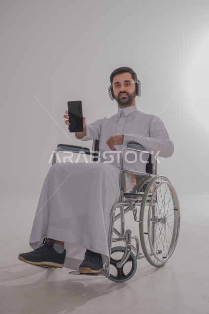 Using modern techniques and advanced technology, portrait of a young Saudi Arabian Gulf man with special needs wearing traditional dress sitting on a wheelchair wearing earphones and holding a mobile phone in his hand, showing a blank black mobile screen, gray background