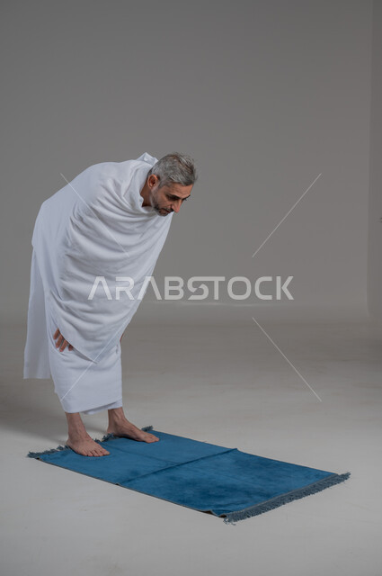 مفهوم الركوع والسجود، قضاء مناسك الحج والعمرة، العبادة والتقرب إلى الله، أداء فرائض الصلاة بأوقاتها، مفهوم أركان الإسلام، بورتريه لرجل عربي خليجي سعودي يرتدي لباس الاحرام يقف على السجادة، موسم الحج 2024، صورة لكامل الجسم، خلفية رمادية
