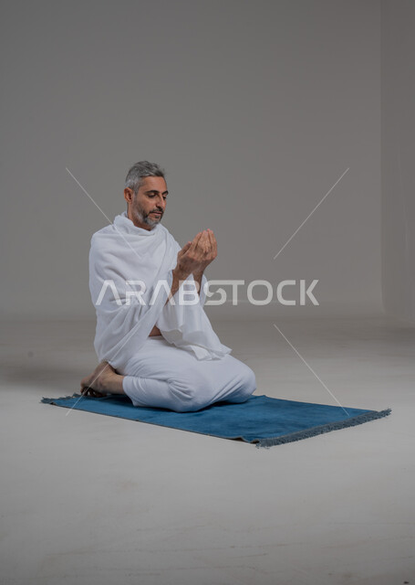 الدعاء و التضرع لرب العباد, مفهوم أركان الإسلام، اداء مناسك الحج والعمرة، العبادة والتقرب الى الله، بورتريه لرجل عربي خليجي سعودي يرتدي لباس الاحرام يجلس على سجادة الصلاة يرفع يديهبايماءات الدعاء، موسم الحج 2024، صورة لكامل الجسم، خلفية رمادية
