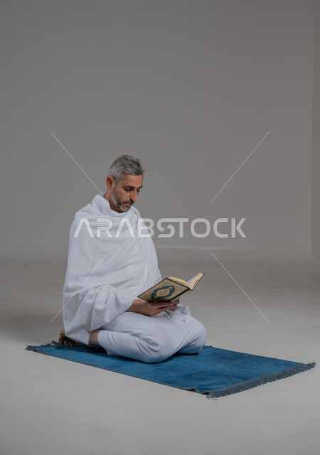 اداء مناسك الحج والعمرة، قراءة وحفظ وتجويد القرآن الكريم, العبادة والتقرب الى الله، بورتريه لرجل عربي خليجي سعودي يرتدي لباس الاحرام يجلس على سجادة الصلاة يحمل بيده المصحف الشريف،تلاوة كتاب الله ، موسم الحج 2024، صورة لكامل الجسم، خلفية رمادية