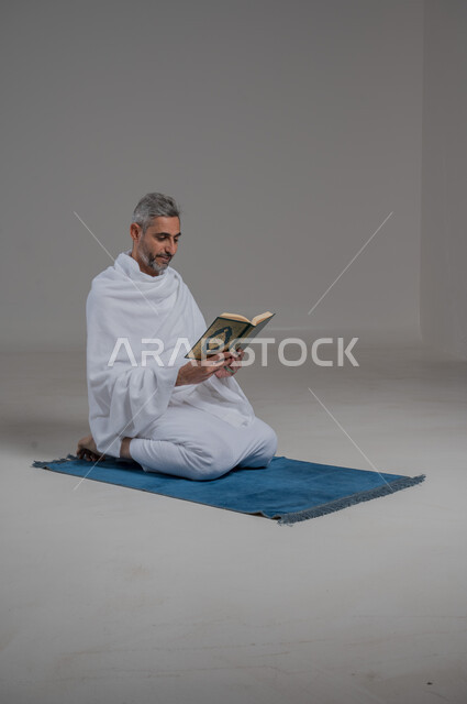 قراءة وحفظ وتجويد القرآن الكريم, العبادة والتقرب الى الله، بورتريه لرجل عربي خليجي سعودي يرتدي لباس الاحرام يجلس على سجادة الصلاة يحمل بيده المصحف الشريف،تلاوة كتاب الله ، موسم الحج 2024، اداء مناسك الحج والعمرة، صورة لكامل الجسم، خلفية رمادية