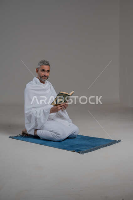 العبادة والتقرب الى الله، قراءة وحفظ وتجويد القرآن الكريم, بورتريه لرجل عربي خليجي سعودي يرتدي لباس الاحرام يجلس على سجادة الصلاة يحمل بيده المصحف الشريف، تلاوة كتاب الله ، موسم الحج 2024، اداء مناسك الحج والعمرة، صورة لكامل الجسم، خلفية رمادية