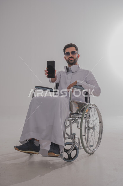 عرض شاشة جوال سوداء فارغة، بورتريه لشاب عربي خليجي سعودي من ذوي الاحتياجات الخاصة يرتدي الثوب التقليدي والنظارات الشمسية يجلس على كرسي متحرك يضع سماعات الأذن، الإمساك بالهاتف المحمول بإيماءات الثقة بالنفس، استخدام التقنيات والتكنولوجيا الحديثة، خلفية رمادية