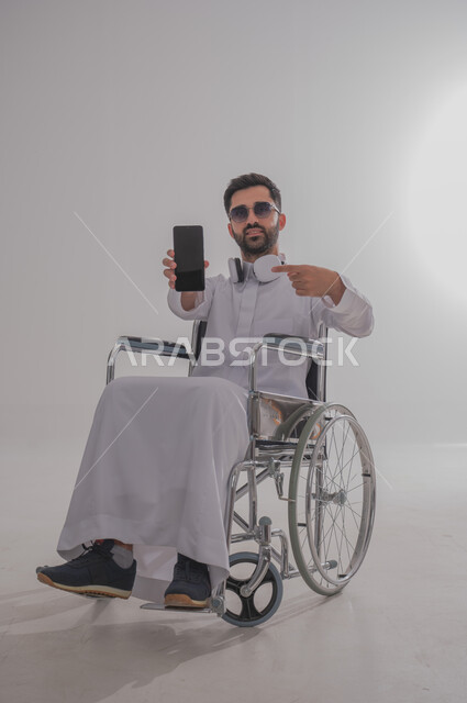 عرض شاشة جوال سوداء فارغة، بورتريه لشاب عربي خليجي سعودي من ذوي الاحتياجات الخاصة يرتدي الثوب التقليدي والنظارات الشمسية يجلس على كرسي متحرك يضع سماعات الأذن، الإمساك بالهاتف المحمول والإشارة إليه بإيماءات الثقة بالنفس، استخدام التقنيات الحديثة، خلفية رمادية