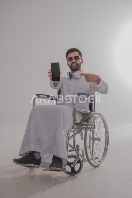 عرض شاشة جوال سوداء فارغة، بورتريه لشاب عربي خليجي سعودي من ذوي الاحتياجات الخاصة يرتدي الثوب التقليدي والنظارات الشمسية يجلس على كرسي متحرك يضع سماعات الأذن، الإمساك بالهاتف المحمول والإشارة إليه بإيماءات الثقة بالنفس، استخدام التقنيات الحديثة، خلفية رمادية
