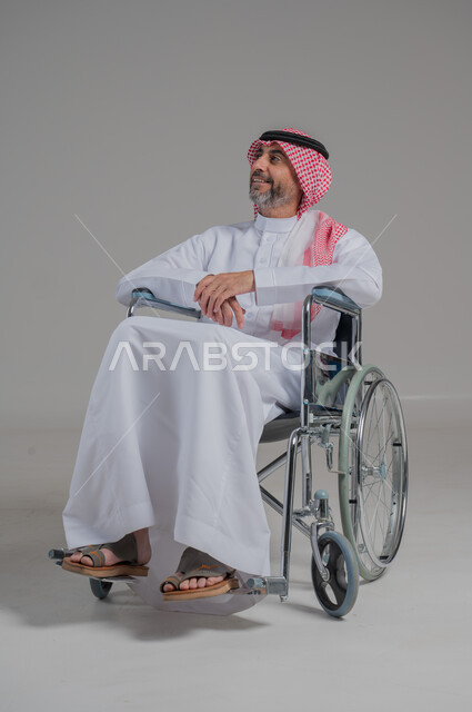 Improving the quality of life of people with motor disabilities, lifestyle of people with disabilities, use of wheelchairs to assist movement and mobility, portrait of a disabled Saudi Arabian Gulf man wearing traditional thobe and shemagh sitting in a wheelchair, full body image, gray background