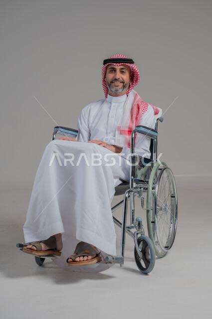 اسلوب حياة ذوي الهمم، استخدام كراسي المعاقين للمساعده على الحركه والتنقل، تحسين جودة حياة ذوي الاعاقه الحركيه، بورتريه لرجل عربي خليجي سعودي مقعد يرتدي الثوب التقليدي والشماغ يجلس على كرسي متحرك، صورة لكامل الجسم، خلفية رمادية