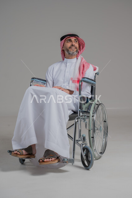 استخدام كراسي المعاقين للمساعده على الحركه والتنقل، اسلوب حياة ذوي الهمم، تحسين جودة حياة ذوي الاعاقه الحركيه، بورتريه لرجل عربي خليجي سعودي مقعد يرتدي الثوب التقليدي والشماغ يجلس على كرسي متحرك، صورة لكامل الجسم، خلفية رمادية
