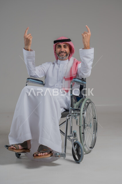 تعيين موقع شيء ما، اسلوب حياة ذوي الهمم، استخدام كراسي المعاقين للمساعده على الحركه والتنقل، بورتريه لرجل عربي خليجي سعودي مقعد يرتدي الثوب التقليدي والشماغ يجلس على كرسي متحرك يشير باصبع السبابة نحو الاعلى، صورة لكامل الجسم، خلفية رمادية