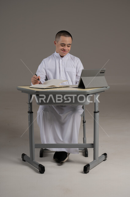 استعمال التابلت لعرض المناهج والمواد التعليمية، دمج ذوي الاحتياجات الخاصة في المدارس العمانية، استخدام التقنيات الحديثة، بورتريه لشاب عربي خليجي عماني مصاب بمتلازمة داون يرتدي الدشداشة يجلس الى طاولة الدراسة يقوم بمراجعه الدروس ، صورة لكامل الجسم، خلفية رمادية